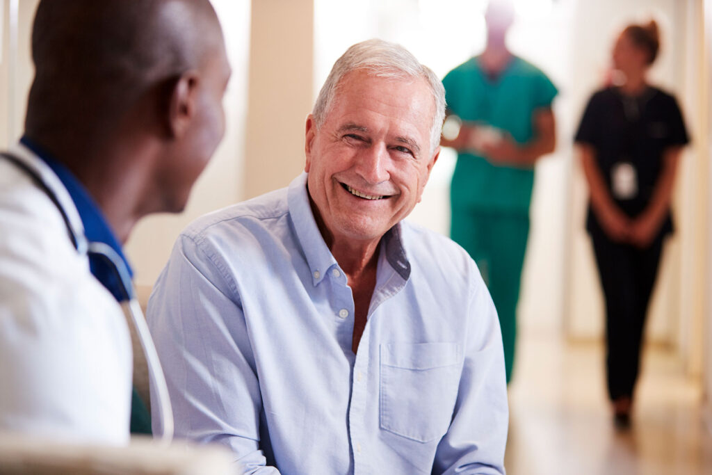 A doctor and patient sitting and smiling with other medical professionals walking and talking in the background,