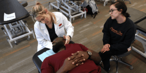 An osteopathic physician manipulates a patient athlete's neck as another watches on.