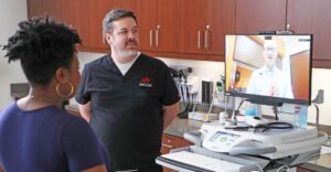 A doctor appears virtually to speak with a nurse and a patient in an examination room.