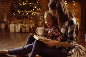 Care Connections Celebrate More A woman and her child sit on the floor reading a book while drinking a hot drink in front of a Christmas tree surrounded by presents.