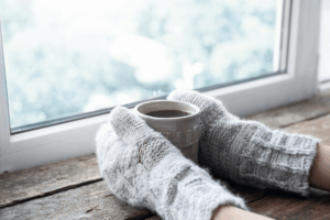 A pair of hands wearing mittens hold a cup of coffee in front of a window.