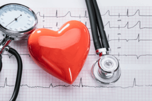 Care Connections Heart_01 A stethoscope. the dial of a blood pressure sensor, and a shiny red heart charm sit atop a piece of graph paper with heart rate monitoring lines drawn of it.