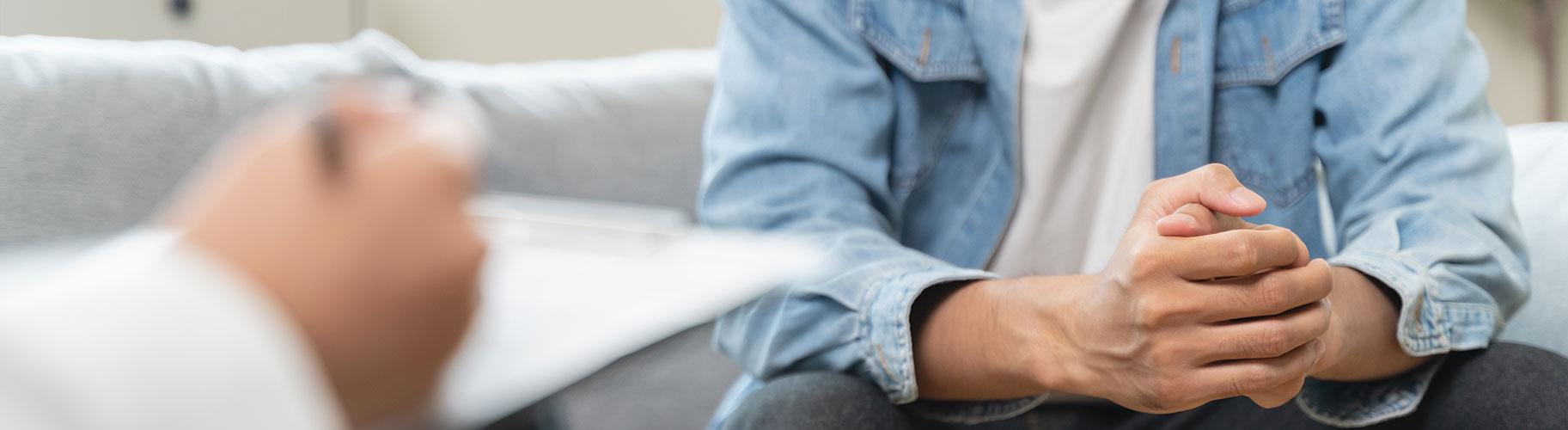 A man sits with hands together across from a clinician taking notes on a clip board.