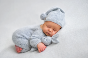 Image of infant for birth defects awareness month A young infant in gray pajamas and a hat balled up sleeping on a gray blanket with a gray stuffed rabbit.