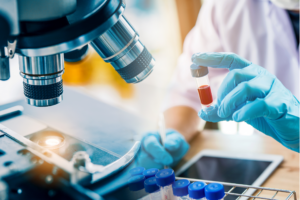 image of clinical researcher A researcher takes notes on a tablet while examining a vial of red liquid next to a microscope.