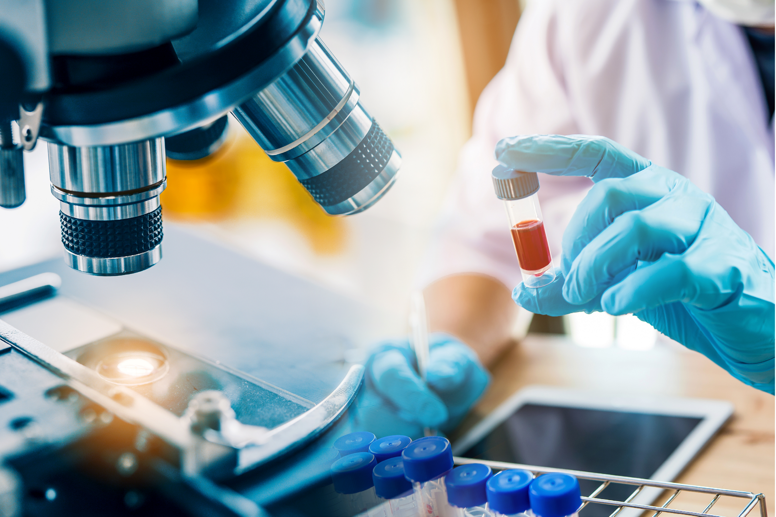 A researcher takes notes on a tablet while examining a vial of red liquid next to a microscope.